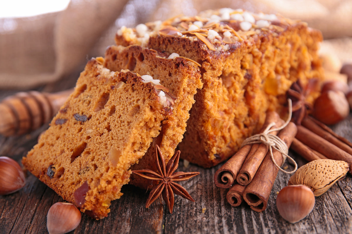 Pain d'épice moelleux garni de fruits secs et d'amandes, accompagné de bâtons de cannelle, d'anis étoilé, de noisettes et d'une cuillère en bois sur un fond rustique en bois.