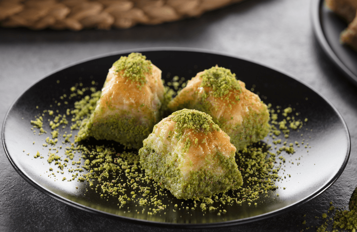 Un plateau de baklava richement doré, empilé en spirale sur un plat décoratif, dans une pièce évoquant une ambiance orientale luxueuse avec des coussins, des tapis ornés et une fenêtre ajourée.