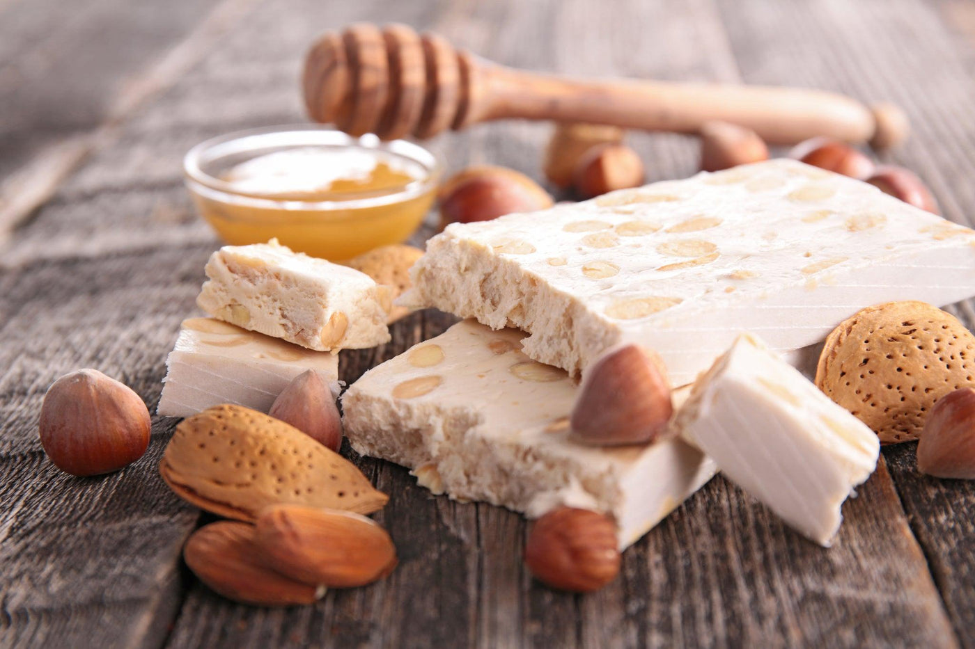 Un plateau de nougat traditionnel parsemé de noisettes et d'amandes, à côté d'un pot de miel et de fruits secs sur une table en bois rustique.